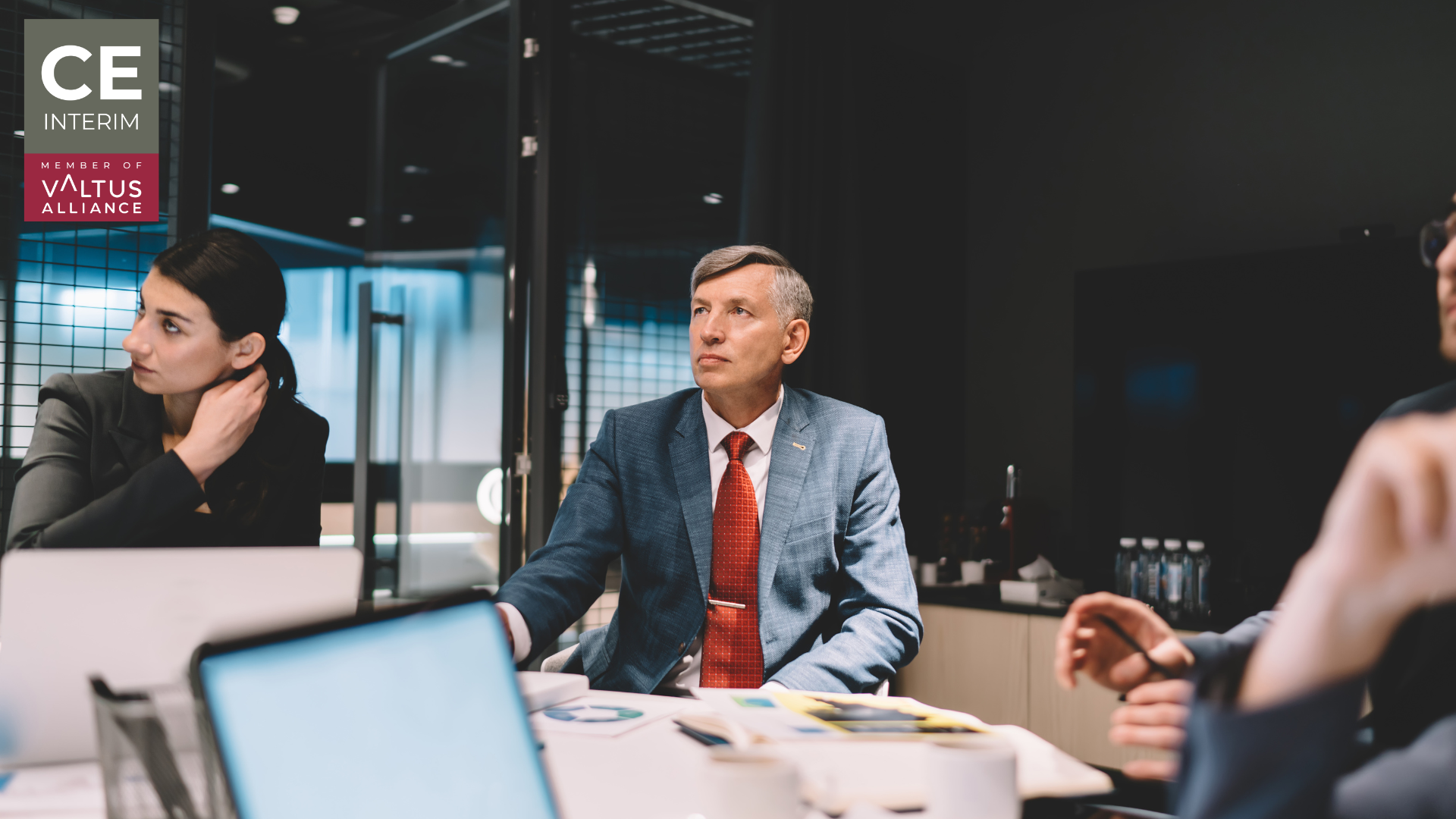A senior interim executive positioned between a Group headquarters boardroom and a manufacturing site.